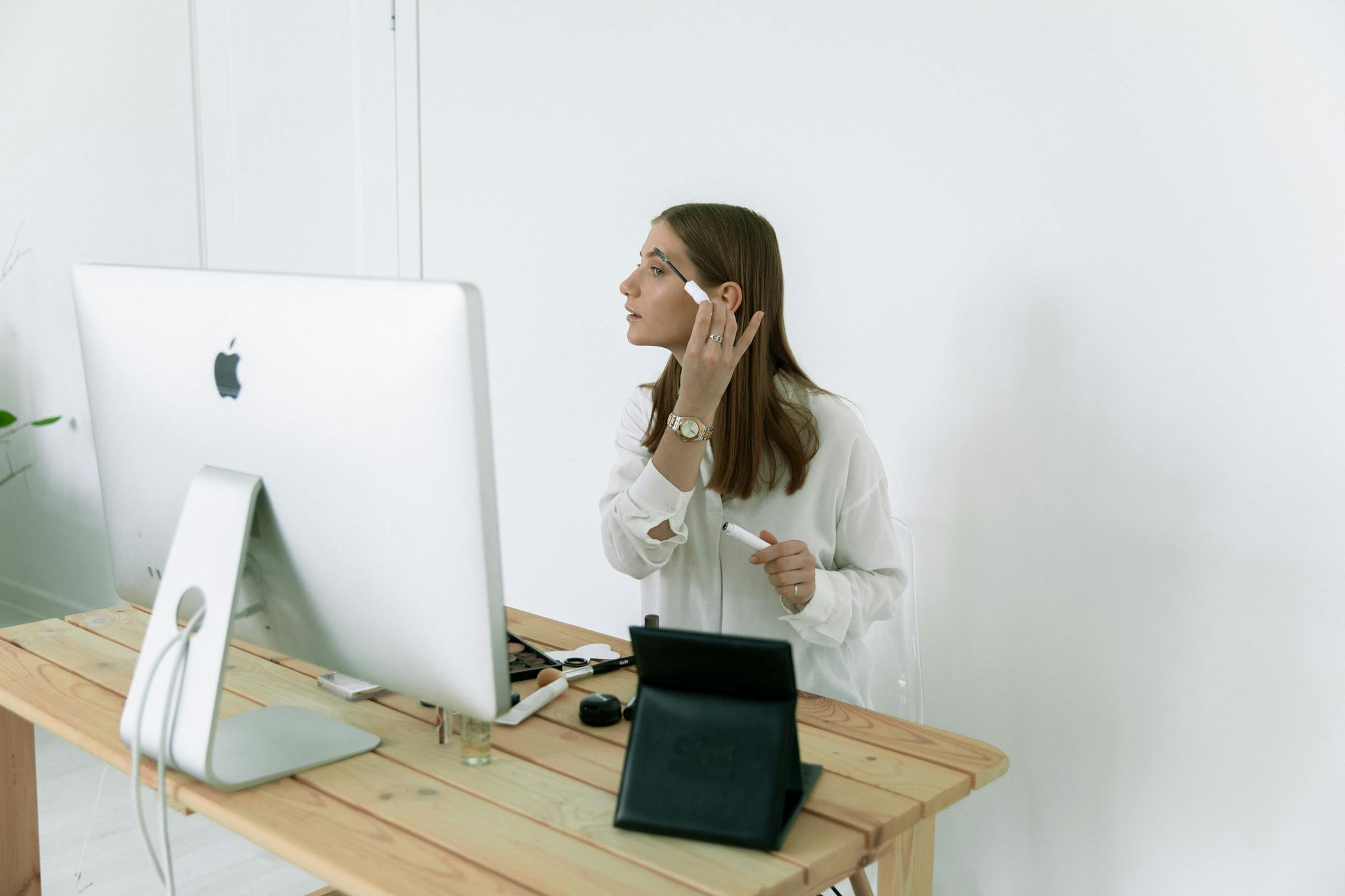 A woman applies makeup while sitting at a desk with a large computer, laptop, and tablet.