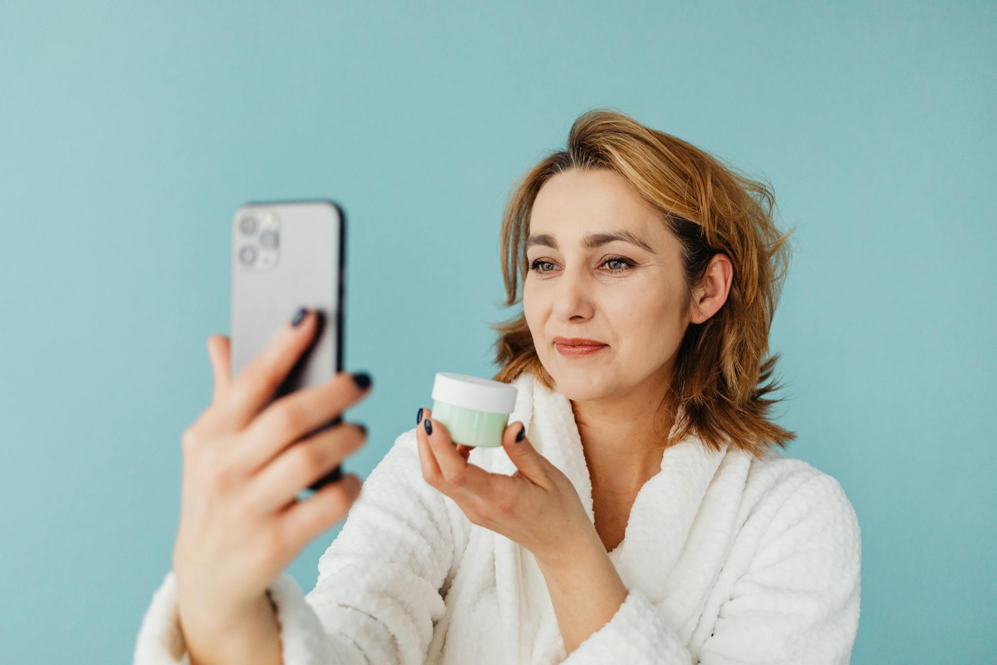 Woman in white robe taking a selfie while holding skincare cream against a blue background.