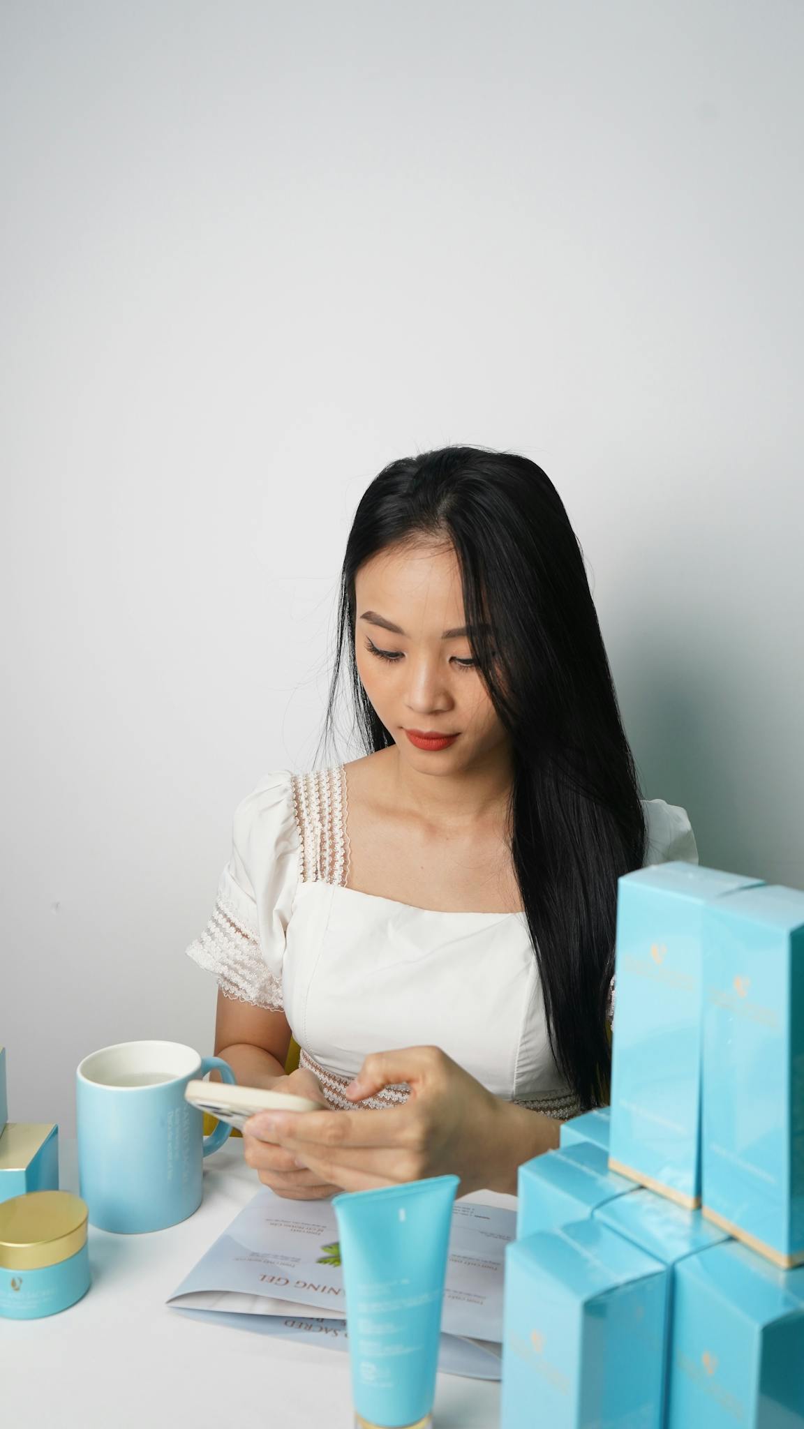 Young woman using smartphone surrounded by skincare products, showcasing modern beauty business.